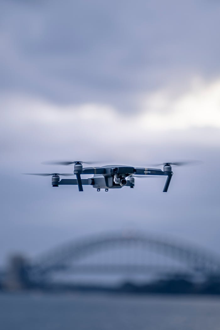 services-02 A drone hovers in front of the iconic Sydney Harbor Bridge against a cloudy sky.